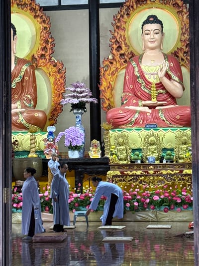 The image captures the interior of a Buddhist temple in Nam Ban, Vietnam. Dominating the scene are two large Buddha statues, one on the left and a more prominent one on the right, both adorned with orange and yellow flame-like halos. The Buddha on the right is seated and displays a gesture of blessing with its right hand while holding a miniature pagoda in its left. Below these statues, a golden altar displays smaller Buddha figures and offerings, including green spheres that resemble fruit and floral arrangements. In the foreground, three women in light blue robes stand or move. One woman on the far left stands facing left, holding a bottle of water. Another woman in the center stands facing right, looking down. A third woman on the right is in motion, bending down towards a small stool. The temple floor is highly reflective, mirroring the statues, flowers, and people. The overall atmosphere suggests a place of worship with ritualistic elements.