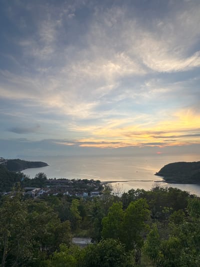 The image depicts a sunset over the ocean in Baan Wang Ta Kien, Thailand. The sky contains layered clouds in hues of grey, blue, and yellow from the sun. The ocean is calm, reflecting the light from the sky, and the land with vegetation and some buildings are on the right side of the picture. A small island is visible.