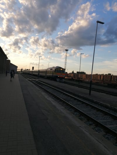 The image depicts a train station scene at sunset, likely in Daugavpils, Latvia. The sky is filled with dramatic clouds, illuminated by the fading sunlight, suggesting an evening time. On the left, the platform is paved with brickwork, leading towards a building that appears to be part of the train station. Several people are visible on the platform, standing or walking, some appearing to be waiting.

The main focus of the image is a train traveling along the tracks. The train is a passenger train with a distinct locomotive at the front, featuring a yellow and red color scheme. It is positioned on a set of railway tracks that run parallel to the platform. Further down the tracks, there are freight cars, indicating a busy operational railway yard. Several lampposts are scattered throughout the scene, some with a modern design. The overall atmosphere seems calm and perhaps a little melancholic, fitting for a sunset scene at a transportation hub. There is no visible text in the image.