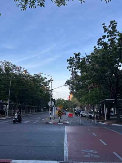 The image depicts a street scene in Tha Khlong Town Municipality, Thailand, during what appears to be late afternoon or early evening. The sky is a pale blue with scattered clouds. Lush green trees line both sides of the road, some with orange-red flowers visible.
In the foreground, a paved road runs through the scene, with a dedicated red-colored bike lane marked on the right side. A person is riding a scooter heading towards the viewer on the main road, passing over a pedestrian crossing marked with white stripes.
Further down the road, there are several parked cars, including a silver SUV. Traffic signs are visible, including a yellow diamond-shaped sign with a black arrow and a stop sign with Thai text. Orange traffic cones and barriers are also present. On the left side of the road, a covered walkway or arcade runs alongside buildings, with more trees and vegetation. Streetlights are mounted on poles along the road. The overall environment suggests an urban or suburban area with ample greenery.