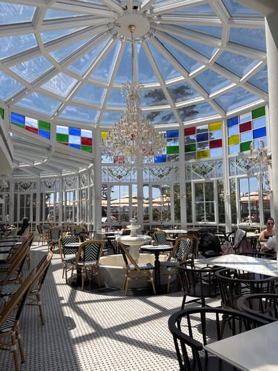 The image depicts an indoor dining area with a distinctive stained-glass ceiling and large windows. The ceiling is a complex geodesic dome structure with panes of clear glass and sections of colored stained glass in red, blue, green, and yellow. A large, ornate crystal chandelier hangs from the center of the ceiling.
The scene is set up for dining, with numerous tables and chairs. The tables have white tops and dark, decorative bases. The chairs are a mix of styles, including some with wicker frames and black and white patterned upholstery, and others with darker, more industrial-looking bases. The floor is tiled with a small, mosaic-like pattern in white and grey. Sunlight streams through the windows and ceiling, casting long shadows across the floor and highlighting the architectural features.
In the background, through the windows, glimpses of an outdoor area with trees and umbrellas can be seen, suggesting it's a pleasant day. There are a few people visible, one woman is seated at a table and appears to be looking towards the camera. A stroller is also visible near one of the tables. The overall atmosphere appears to be upscale and airy, likely a restaurant or cafe. The time of day is clearly daytime due to the strong sunlight and shadows. The location context suggests this scene is in Üsküdar, Turkey. There is no visible text in the image.