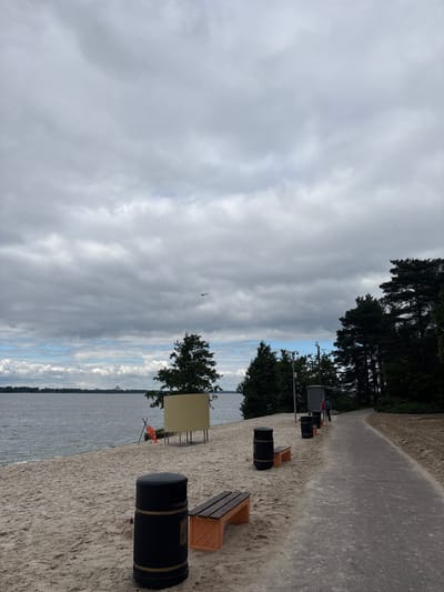 The image depicts a cloudy day at the beach in Sestroretsk, Russia. On the left, a sandy beach runs along a body of water. There are several trash cans and benches on the beach, as well as a changing booth. A life preserver is visible on a stand near the water. In the distance, a line of trees marks the horizon. On the right, a paved path runs along the beach, bordered by trees. Two individuals are seen walking on the path. The sky is overcast with scattered clouds.