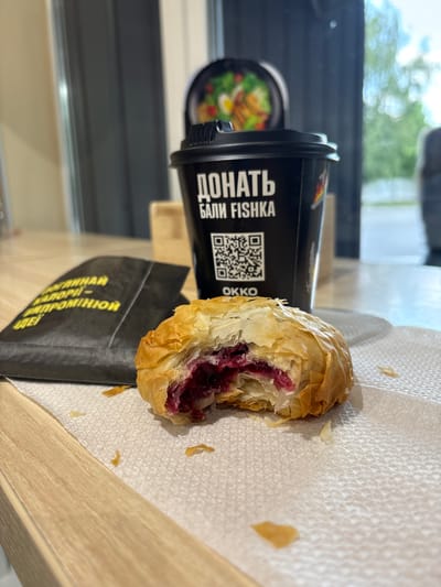 A close-up, low-angle shot shows a partially eaten pastry with a dark berry filling on a white paper napkin. The pastry is golden brown and flaky. To the left of the pastry is a black paper bag with yellow text. Behind the pastry and bag is a black paper cup with white text and a QR code. The cup appears to be for coffee. In the background, out of focus, is a window with a view of greenery and a bright sky. The setting appears to be an indoor cafe or food establishment, possibly in Bila Krynytsia, Ukraine, given the context of the text on the cup and bag. The lighting is natural, coming from the right, creating subtle shadows on the table.
