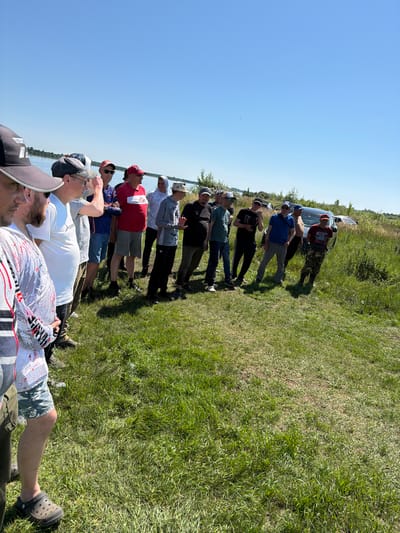 In Tarasovo, Russia, a group of approximately 15 men stand in a grassy field near a body of water under a clear blue sky. The men are of various ages and are dressed in casual attire, including t-shirts, shorts, and hats. Some are wearing baseball caps, while others have on bucket hats or headscarves. The men appear to be gathered for an informal event or activity. Several vehicles, including a dark SUV and a white car, are parked in the background behind the group. The environment is characterized by lush green grass, with some patches of dry earth. In the distance, trees line the shore of the water. The overall scene depicts a warm, sunny day outdoors.