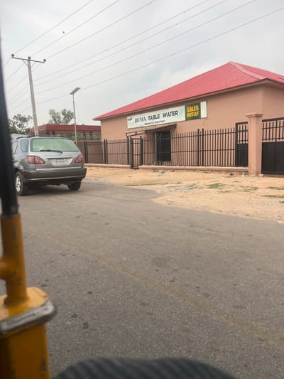 The image shows a building in Gulum, Nigeria, identified by a sign as "BUMA TABLE WATER SALES OUTLET." The building is a single-story structure with a red roof and a light beige exterior. A black metal fence surrounds the property. A silver SUV is parked on the asphalt road in front of the building. Overhead, there are power lines supported by utility poles. The sky is overcast. The image appears to have been taken from a moving vehicle, evidenced by the blurred yellow object at the bottom.