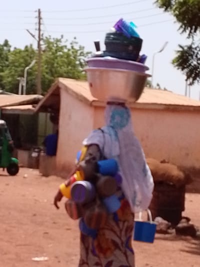 This image captures a bright, sunny street scene in Dungu, Ghana, depicting a person engaged in daily commerce or transport. The main subject is a person, likely a woman, seen from the back, carrying a substantial load of various items.

She balances a large, shiny metallic bowl on her head, which is topped with several smaller containers including a purple bowl, a black basket-like item filled with bright blue and purple objects, and other plastic articles. Her head is covered with a white fabric featuring light blue patterns. Strapped to her front are numerous colorful plastic containers—yellow, blue, purple, and red—suggesting she is a vendor transporting goods for sale. A blue shopping bag is visible hanging from her left side. She wears a long, patterned dress or skirt with shades of brown, orange, and blue.

The ground is unpaved, consisting of reddish-brown dirt. In the background, simple, light-colored buildings with brown roofs are visible, characteristic of local architecture. Green trees are interspersed throughout the scene, and a utility pole with wires along with a street light stand against the bright, clear sky. To the left, part of a green three-wheeled vehicle is visible, and another person in a blue shirt can be faintly seen inside one of the background structures. To the right, a large, dark, possibly woven storage object rests on the ground.

The scene illustrates the common practice of carrying goods on the head in the region, a method of transportation for vendors and individuals alike. The time of day is clearly daylight, under a bright sky. No visible text is discernible in the image.