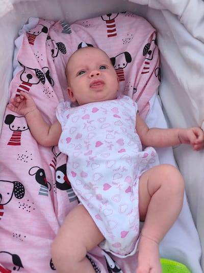 This is a top-down, eye-level shot of a baby lying in a stroller, situated in Stryków, Poland. The baby, likely an infant, has fair skin and blue eyes, and is looking upwards with its mouth slightly open and tongue sticking out. The baby is wearing a white onesie adorned with a pattern of pink hearts and the words "only you" repeated in cursive. The stroller is lined with a pink blanket featuring a playful pattern of cartoon dogs. The baby's right arm is resting on the blanket, while the left arm is extended slightly to the side. The lighting suggests it is daytime, and the outdoor setting is implied by the stroller and the natural light. The overall mood is one of gentle observation, capturing a candid moment of the baby's alertness. Visible text includes "only you", "only yo", "onl", and "Onky".