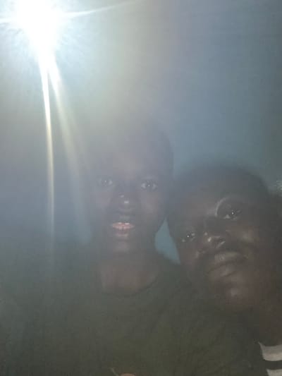 Two individuals are visible in the image. They appear to be in close proximity, possibly posing for a photograph. The environment is dimly lit with a bright light source on the left obscuring details. The location is given as Epe, Nigeria, but the image does not provide visual confirmation of the city or specific details.