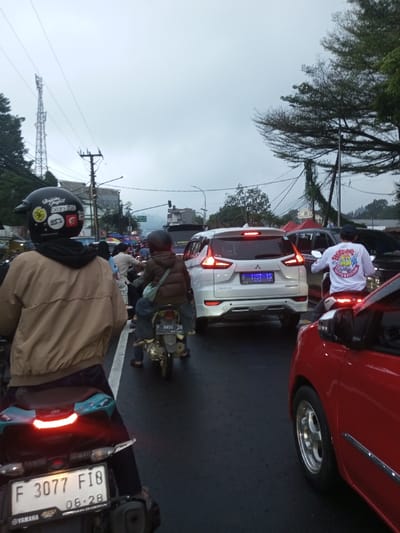 The image depicts a crowded street scene in Dawathang_Dorjibi_ Kashingtsawa, Bhutan, under an overcast sky. The road surface appears damp. Multiple vehicles, primarily motorcycles and cars, are visible in apparent traffic congestion.

In the immediate foreground, facing away from the viewer, is a person on a black Yamaha motorcycle. The rider wears a black helmet adorned with several stickers, including a Turkish flag and text such as "DODTIS," a black hooded garment, and a light brown bomber jacket. The motorcycle's license plate, partially visible, reads "F 3077 FI8" with "06-28" below. To the right of this motorcycle, the right side of a red car is visible, showing a silver multi-spoke wheel and an illuminated brake light.

Further into the scene, directly in front of the foreground motorcycle, a person rides a smaller motorcycle or scooter. This rider wears a dark helmet, a brown puffer jacket, blue jeans, and carries a light blue sling bag. Their license plate reads "F 5850 ZW" with "09-27" below. Immediately to the right of this motorcycle is a white SUV, identified as a Mitsubishi Xpander, with its brake lights illuminated. Its rear license plate reads "F 1157 YS." To the right of the white SUV, another person on a motorcycle is visible from behind, wearing a white long-sleeved shirt with colorful graphics and text, including "BISNIS KECIL KU" and "KOPI & CAFE," and a dark cap.

The left side of the street features various commercial structures or market stalls, with awnings visible. A tall, lattice-structured communication tower stands prominently in the background on the left, alongside a utility pole with numerous overhead wires. On the right side of the street, large green trees line the road, with power lines running through them. A red tent or awning is partially visible in the distant background on the right. Additional vehicles and individuals are visible further down the street, indicating ongoing urban activity.