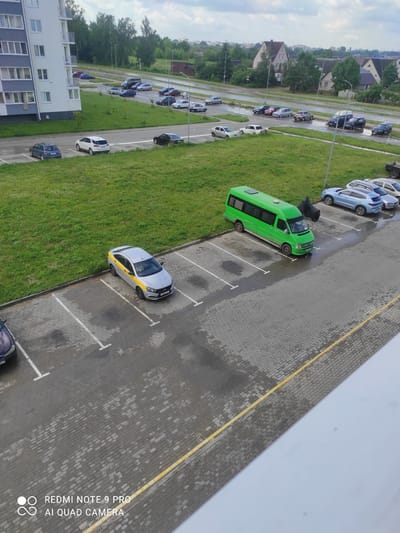 The image shows a parking lot in front of a multi-story building, likely in the city of Maladzyechna, Belarus. The parking lot is paved with grey interlocking bricks, and white lines demarcate parking spaces. Several vehicles are present, including a silver sedan with a yellow stripe, a green minibus, and various other cars parked in the lot and on the adjacent road. The building appears to be an apartment complex with a modern facade. The surrounding environment includes a grassy area, trees, and other buildings in the background, suggesting a residential or mixed-use urban area. The sky is overcast, and there are signs of wetness on the ground, indicating recent rain. The image is captured from an elevated perspective, possibly from a window or balcony.