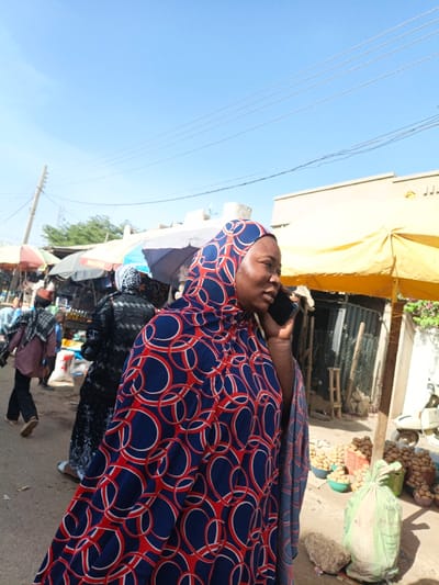 The image depicts an outdoor market scene in Kawaji, Nigeria, under a clear blue sky. In the foreground, a woman faces right, holding a black smartphone to her right ear, engaged in a call. She wears a blue head covering and garment patterned with red and white concentric circles. To her left, in the middle ground, other individuals are visible, including one person walking away wearing a checkered head covering and another in a dark patterned dress. Multiple market stalls with various colored umbrellas (white, red, dark) line what appears to be a street or pathway. Utility poles with overhead wires are visible against the sky on the left. To the right of the primary subject, a market stall operates under a large yellow umbrella. Piles of light-colored, round produce, possibly potatoes, are arranged on the ground, some contained within green bowls and a red basket. A large light green sack rests on the ground next to a rock. A white scooter or motorcycle is partially visible on the far right. A light-colored building and a corrugated metal structure are present in the background on the right. The scene is brightly lit by natural sunlight.