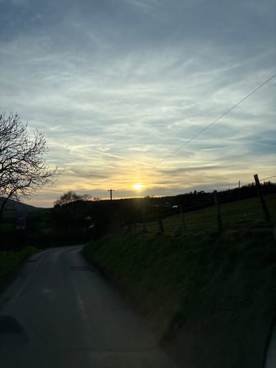 The image depicts a rural landscape scene at dusk or dawn in Shelve, United Kingdom. A dark asphalt road curves from the lower left foreground towards the mid-distance. To the left, a bare deciduous tree partially obscures a white triangular road sign with a red border. Rolling green hills are visible in the background on the left. On the right, a steep, dark green grassy bank is present, bordered by a wooden post and wire fence. Beyond the fence, a field leads to a treeline silhouetted against the horizon. A dark utility pole with cross arms stands silhouetted slightly left of the low-lying sun. The sky transitions from an orange-yellow glow at the horizon to a lighter blue, containing wispy white clouds and two visible aircraft contrails. A single dark wire extends diagonally across the upper sky. No individuals, animals, or vehicles are visible in the frame.
