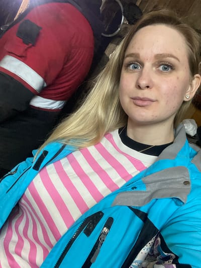 A young Caucasian woman with blonde hair and blue eyes is pictured from the chest up, looking directly at the camera. She is wearing a pink and white striped long-sleeved shirt under a bright blue ski jacket with gray accents. She has a small diamond stud earring in her right ear. In the background, a person in a red jacket is partially visible, and the environment appears to be indoors, possibly a workshop or storage area, with wooden beams and metal objects. The scene does not provide any specific indicators of its location within Kauchuk Stantsiya, Russia, beyond what is stated.