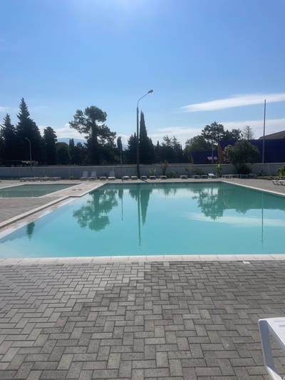This image captures a serene outdoor scene, likely in Giachrypsh, Abkhazia, featuring a swimming pool area. The primary focus is a large, rectangular swimming pool filled with clear, turquoise water, reflecting the bright blue sky and surrounding trees. The pool is surrounded by a paved area made of grey interlocking rectangular bricks arranged in a herringbone pattern. To the left of the main pool, a smaller, shallow pool or wading area is visible, also lined with the same grey brickwork.
Several white, empty lounge chairs are neatly arranged around the pools, suggesting a well-maintained recreational facility. In the background, a line of lush green trees, including tall cypress and pine varieties, forms a natural backdrop. Beyond the trees, hints of buildings and a mountain range are discernible under the clear, sunny sky. The overall atmosphere is peaceful and inviting, indicative of a resort or leisure complex. There are no people visible in the image.
