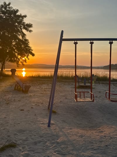 This media file depicts an outdoor playground scene at sunset, likely in Berezhany, Ukraine. The primary subjects are playground equipment, including swings and a seesaw, set against a backdrop of a serene body of water and a distant treeline. The low sun casts a warm, golden glow across the scene, illuminating the sandy ground and highlighting the textures of footprints in the sand.

The image captures a peaceful, almost tranquil moment as the day draws to a close. The sky is a gradient of soft blues and warm oranges, indicative of sunset. The water reflects the vibrant colors of the sky, creating a shimmering effect. The playground equipment, though stationary, evokes a sense of past activity and the anticipation of future play. The presence of a single tree in the foreground adds a touch of natural beauty to the man-made environment.

Notable details include the dramatic lighting of the sunset, which creates long shadows and a warm atmosphere. The location cues, such as the type of vegetation and the overall landscape, are consistent with a natural setting near water, potentially a lake or river. The emotion conveyed is one of calm, beauty, and a hint of nostalgia associated with childhood memories of playgrounds. There is no visible text in the media file.