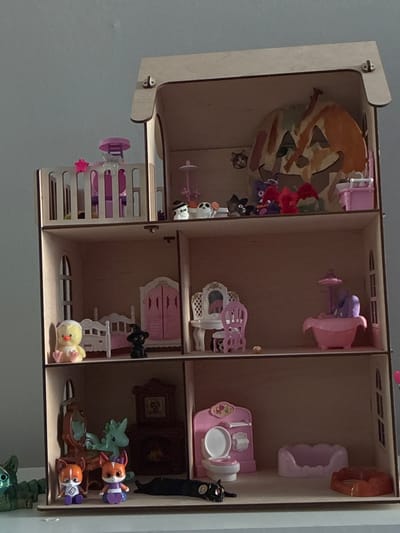 The image shows a dollhouse filled with miniature furniture and figures. The dollhouse is made of light-colored wood and is divided into several rooms.
In the top left room, a stylized image of a fox or dog is painted on the back wall. There are also some small decorative flowers and figures in this room.
The top middle room contains a pink and purple toy bathtub or jacuzzi and a small pink chair.
The top right room has a light purple, shell-shaped object and a pink dollhouse toilet with a sink and a small decorative plate. A black cat figurine is next to the toilet.
The middle left section has a pink crib or bed with a small pink star attached.
The middle middle room features a pink double door and a white ladder. There are also several small black cat figurines and a yellow chick figurine.
The middle right room has a miniature framed picture on the wall, and below it, a dollhouse vanity with a mirror. In the bottom right section of the dollhouse, there are several animal figurines, including what appear to be dog-like figures in purple outfits, and a light green dragon-like figure. There is also a brown furniture piece, possibly a bed or couch, with these figures.
The overall scene is a collection of toys and miniature items arranged within a dollhouse structure. There is no indication of specific time of day, weather, or any discernible emotions. No text is visible in the image. The location context provided is "Гаранскі сельскі Савет, Belarus," which suggests the setting is a rural area in Belarus.