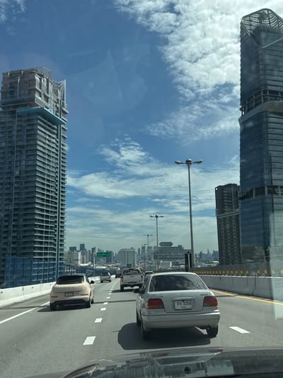 The image depicts a forward-facing view from a vehicle traveling on an elevated highway in Song Khanong Subdistrict, Thailand. The highway is flanked by concrete barriers and has multiple lanes marked with white dashed lines and solid yellow lines. The sky is blue with scattered white clouds.

Dominating the left side of the frame is a tall, modern high-rise building under construction, characterized by its numerous floors and visible structural elements. To the right, another sleek, modern skyscraper with a distinctive angular top and glass facade is prominent. In the distance, a cityscape with numerous other buildings is visible.

Several vehicles are present on the highway. In the foreground, the hood and windshield of the reporting vehicle are visible. Ahead, a light-colored SUV is in the left lane, and a silver sedan with a license plate that reads "กง 2258" is in the right lane. Further ahead, a white pickup truck and another vehicle are also on the highway. Traffic lights and street lamps are visible along the elevated road. A blue construction barrier is present along the left edge of the highway.