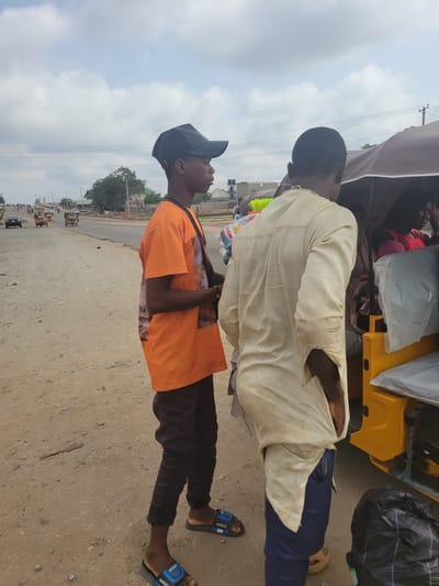 Two men are standing on a dirt shoulder of a road in Kwamba, Nigeria. One man is wearing a light yellow shirt and blue pants and has black sandals on his feet. The other man is wearing an orange shirt, dark-colored pants, and black and blue sandals. He has a black baseball cap on his head. They are standing next to a yellow vehicle that appears to be a commercial vehicle with a canvas top. There is baggage on top of the vehicle. The road has light traffic consisting of other commercial vehicles, cars, and motorcycles. The sky is overcast.