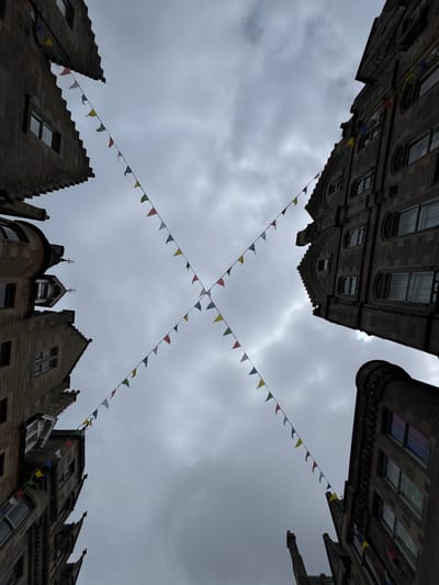 This is a low-angle, upward-facing photograph of a street in Edinburgh, Scotland. Four buildings, each with stone facades and multiple windows, are visible at the edges of the frame, creating a sense of being in a narrow, enclosed urban space. A string of colorful triangular pennant flags crisscrosses the sky, forming an 'X' shape in the center of the image. The sky is overcast and appears to be a muted grey, with some lighter patches suggesting cloud breaks. The buildings exhibit architectural details such as arched windows, carved stonework, and pointed gables, consistent with the historic architecture found in the city center of Edinburgh. There are no people visible in the image. The overall atmosphere is somewhat somber due to the cloudy sky, but the colorful flags add a festive touch.