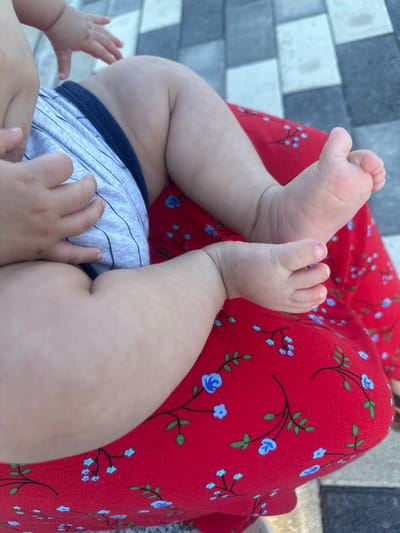 The image shows a close-up of a baby's legs and feet, resting on a red fabric with a blue floral pattern. The baby is wearing light gray shorts with dark blue trim. The baby's feet are small and delicate, with visible toes and fingernails. One foot is positioned above the other, and the baby's legs are bent at the knees. The background consists of a gray and white tiled surface, possibly a patio or walkway. The scene appears to be outdoors, as suggested by the lighting and the texture of the ground. The overall impression is one of tenderness and domesticity. There is no visible text in the image. The location context provided is Manas, Kyrgyzstan, which adds a geographical element to the scene, though it is not directly depicted in the image itself. The time of day and weather are not discernible. The emotion conveyed is likely one of peacefulness or relaxation, as the baby appears to be resting.