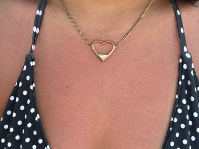 A close-up, overhead shot shows a woman's chest and neck, adorned with a delicate gold necklace in the shape of a heart. The necklace is set against her skin, which appears to have a slight tan, possibly from sun exposure. The woman is wearing a black and white polka dot bikini top, the straps of which are visible on either side of the frame, framing her décolletage. The fabric of the bikini is slightly textured, with the white dots standing out against the black background. The heart pendant of the necklace is small and appears to be made of gold, with a small, possibly diamond, accent at its center. Inside the heart, the word "PRADA" is visible in white text, indicating the brand of the jewelry. The background is her skin, which shows a subtle texture, and no other discernible background elements are present. The lighting suggests it is daytime, and the overall impression is one of a relaxed, possibly beach or poolside setting, given the bikini attire. The image is focused on the details of the jewelry and the bikini, highlighting the personal style of the wearer.