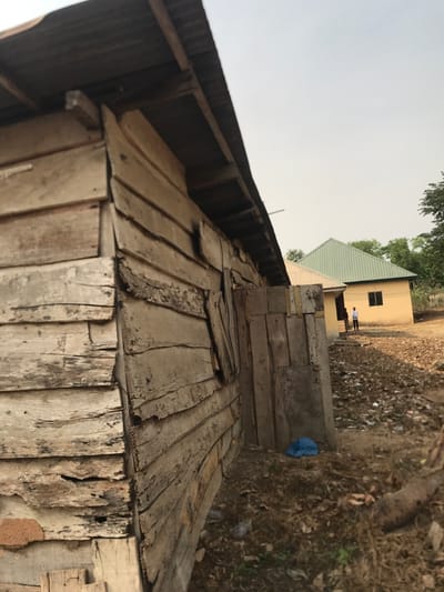 The image depicts an outdoor scene, likely in a rural or semi-urban setting, consistent with Umudike, Nigeria. The foreground is dominated by a weathered wooden structure, possibly a wall or shed, constructed from rough, uneven wooden planks. These planks show significant signs of age, wear, and discoloration, with various shades of grey and exposed grain. The top of this structure appears to have corrugated metal sheeting.
Beyond the wooden structure, a path of dirt and dry ground leads towards a light yellow or tan-colored building in the mid-ground. This building features a sloped, light green corrugated metal roof and a visible rectangular window. Two indistinct figures are standing near this building; one individual is more clearly visible wearing a light-colored shirt. The ground surrounding the pathway and buildings is characterized by dry grass, patches of earth, and scattered debris, including a blue bag or tarp near the wooden structure.
The sky in the background is a uniform, pale grey, suggesting an overcast or hazy day. The lighting is diffused, indicating either midday or late afternoon without direct sunlight. There is no visible text in the image, and no specific activities or emotions are clearly discernible from the figures.
