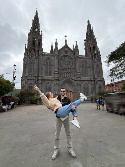 The image captures a daytime scene in a paved public square in Arucas, Gran Canaria, Spain, with the Parroquia de San Juan Bautista (Church of San Juan Bautista) prominent in the background under an overcast sky. In the foreground, a male, estimated to be in his 20s-30s, wearing a black hooded sweatshirt, light grey trousers, and sunglasses, stands holding a female, also estimated to be in her 20s-30s, horizontally in his arms. The female, dressed in a beige fluffy sweater and light blue jeans, has her right arm raised in a peace sign gesture and is looking at the male. The male is looking at the female. The church behind them is a dark grey neo-Gothic structure with two prominent spires, a large central rose window, and multiple ornate arches and windows. To the far left, a group of individuals, including children, are visible near green bushes and a multi-headed lamppost. To the right, two adult individuals stand in the mid-ground, with a stack of wooden pallets and a building with red and white facades further in the background.