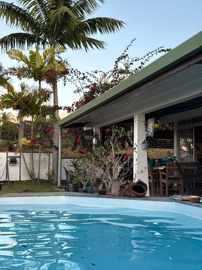The image depicts an outdoor residential scene in Māhina, France, featuring a swimming pool and a covered patio area with lush vegetation. The foreground is dominated by a light blue, irregularly shaped swimming pool with visible ripples on its surface and a few small dark specks of debris. The pool's edge, a light-colored coping, is visible along the right and top edges of the water.
Behind the pool, to the right, is a covered patio or veranda with a light green corrugated metal roof and a white soffit. The patio is supported by a thick white pillar. Beneath the covered area, various objects are present: several potted plants, including a large, somewhat gnarled plant with sparse pink blooms, and another plant in a hanging basket. A large, dark brown, urn-shaped clay pot lies on its side on the patio floor near the pool edge. Further inside the patio area, a wooden dining table with several chairs is visible, along with a dark-colored wicker-style chair. Other miscellaneous items are scattered, including what appears to be a small blue watering can hanging from the pillar and a colorful circular object on the ground near the wicker chair.
To the left and rear of the patio, dense tropical foliage is present. A prominent feature is a collection of palm trees, some tall with visible trunks and others shorter. A white horizontal fence with dark vertical supports runs along the mid-ground, behind which more green vegetation is visible. Bright red bougainvillea-like flowers are draped over the patio roof and bloom near the fence line. A green hose reel is mounted on the fence to the far left. The sky above is clear and light blue, indicating daylight conditions. No individuals are present in the scene.