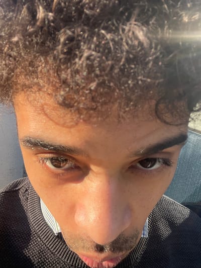 A close-up, high-angle portrait shows a single individual, identified as being in Montigny-le-Bretonneux, France. The subject is a male with medium-dark skin, dark brown eyes, and dark, tightly curled hair. He has defined eyebrows and a faint mustache. He is wearing a dark, possibly black or charcoal, knitted crew-neck sweater over a light-colored, possibly striped, collared shirt, visible at the neckline. Strong, direct sunlight illuminates the face, creating bright highlights on the forehead, nose, and cheeks, and casting shadows that emphasize facial contours. A lens flare is present in the upper right portion of the image. The subject is looking directly into the camera. The background is out of focus, with indistinct light and dark areas suggesting an outdoor environment.
