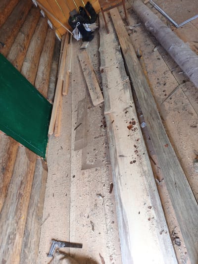 The image depicts the interior of a rustic structure, possibly a shed or unfinished room, located in Islambaevo, Russia. The walls are constructed from rough-hewn logs. The floor is made of wooden planks and is covered in sawdust and wood shavings, indicating recent construction or woodworking activity. Several lengths of lumber are scattered across the floor, with one particularly large, light-colored plank dominating the center of the frame. To the left, a section of wall is painted a vibrant green. In the upper right quadrant, a large roll of grey material, possibly insulation or roofing felt, is visible. Near the top of the image, a pair of black boots and some plastic bags are piled. In the lower left corner, a small hand saw rests on a piece of burlap or sacking. The overall impression is one of a work-in-progress or storage area for building materials.