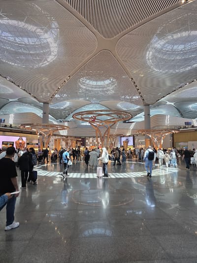 The image captures an interior scene at Istanbul Airport, located in Arnavutköy, Turkey. The perspective is tilted, emphasizing the architectural details of the ceiling. The ceiling is constructed with a white, linear pattern, creating a latticework effect. People are seen walking along a brightly lit corridor lined with shops and stores. Individuals are focused on their own tasks, such as walking and shopping. A circular, decorative structure is a focal point, positioned in the center. The floor is a polished, light-colored material that reflects the ceiling’s design and the surrounding light.