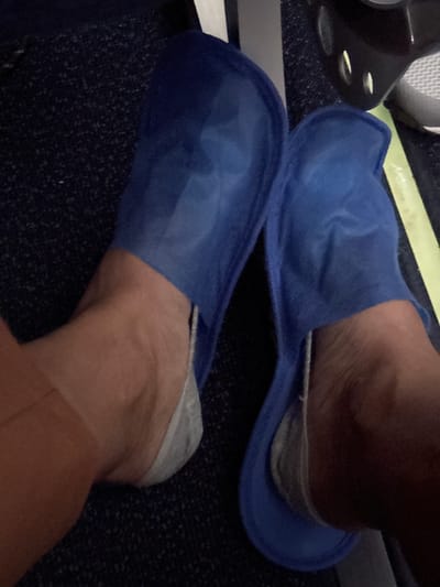 The image shows a close-up view of a person's feet wearing blue disposable slippers and grey socks. The feet are resting on a dark, textured surface, possibly airplane carpeting. A portion of a black seat with metallic accents and a bright yellow stripe is visible in the upper right corner. The lighting is dim, suggesting an indoor environment. The scene is devoid of any identifying features that would place it specifically in Malye Kabany, Russia.