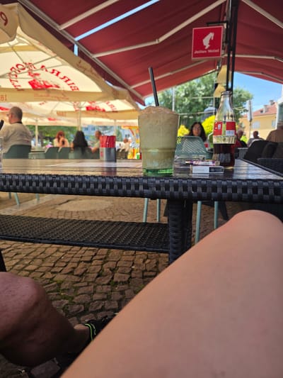 An outdoor cafe scene shows a close-up of a table with a drink, a bottle of Coca-Cola, and a cigarette pack of Sobranie Black Russian cigarettes. In the background, people are seated at tables under red and white awnings. The ground is paved with cobblestones. The scene appears to be set in Dupnitsa, Bulgaria, as suggested by the "Julius Meinl" signage, a company with a presence in Bulgaria.