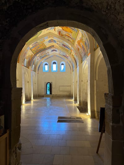 The image shows the interior of a religious building, likely a church, with a stone archway in the foreground. The ceiling is adorned with colorful religious frescoes depicting scenes with figures. Several arched windows are visible, letting in natural light. The floor is tiled and reflects the light. The architecture features stone elements and arched doorways. The lighting is dim, with artificial lights illuminating the interior, creating dramatic shadows and highlights. The overall impression is one of solemnity and historical significance. No specific activity or event is clearly depicted, suggesting a quiet or contemplative moment within the space. The setting appears to be Dzegvi, Georgia, a location known for its historical religious sites. The frescoes suggest a rich cultural and spiritual heritage.