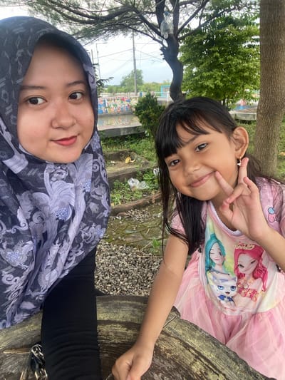 Two individuals are captured in a selfie-style image, presumably taken outdoors. The younger individual, a child, is positioned in the upper portion of the frame, wearing a pink dress and making a peace sign with their hand. The other individual, an adult woman wearing a hijab, is situated at the bottom of the frame. Both individuals appear to be smiling. The setting appears to be in the city of Kalitengah, Indonesia, as evidenced by the vegetation, trees, and infrastructure in the background. The ground consists of small gravel or stones. A wooden surface is also visible, suggesting the individuals are sitting or lying on a bench or similar structure.