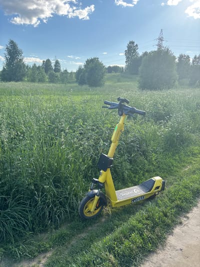 A bright yellow electric scooter is parked on a dirt path next to a grassy field. The scooter has black handlebars and a black deck with a pattern on it. The text "HIMO CO" and "HM549X" are visible on the scooter. The background is a field of tall green grass with some lighter colored wildflowers scattered throughout. In the distance, there are trees and a tall power line tower under a bright blue sky with scattered white clouds. The sun is shining brightly, casting shadows on the grass and path, suggesting it is daytime. The scene appears to be in a rural or semi-rural area, possibly a park or undeveloped land, consistent with the location context of Koltushi, Russia. There are no people visible in the image, and the scooter is stationary, implying it has been parked. The weather appears to be warm and sunny.