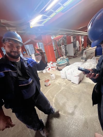 An indoor, industrial scene in Zlatitsa, Bulgaria, is depicted from an angled perspective, with the original image appearing rotated counter-clockwise. In the foreground, a person wearing a blue hard hat, safety glasses, and dark blue work overalls with blue trim, looks towards the camera. They have a beard and are holding a yellow-tipped brush or tool in their right hand.

To their right, on the concrete floor, are several open paint cans, some with brushes inside, and a small white plastic container. Further back, a stack of white, plastic-wrapped bags is visible, many displaying a red 'X' hazard symbol in a square. Nearby, there is a blue and grey industrial machine, possibly a floor grinder or polisher, and other technical equipment and machinery in the background. Another person's hands and lower arm are visible towards the upper right of the frame, holding a dark object, possibly a smartphone or tool.

The background features red structural beams, metal scaffolding or shelving, and bright fluorescent lighting fixtures, causing some lens flare. The concrete floor appears somewhat dusty or covered with fine particulate matter, potentially from the materials in the bags or general industrial activity. The overall setting is a busy, functional workspace, likely involved in construction, renovation, or material processing. The time of day is not discernible due to the dominant artificial lighting.