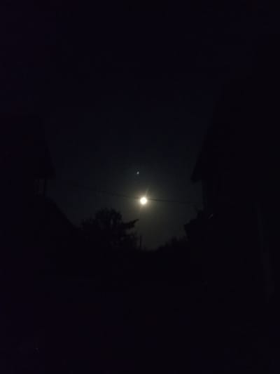 The image depicts a dark, nighttime scene with a bright moon in the sky. Silhouetted against the moonlight are the outlines of two buildings on either side of the frame, suggesting a residential or urban setting. In the center of the image, there is a silhouette of a tree or cluster of trees. A faint, illuminated line, possibly a power line or string of lights, stretches horizontally across the frame, passing near the moon. There are no visible people or text. The overall atmosphere is one of quiet darkness, with the moon being the dominant source of light. The location context provided is Pochaiv, Ukraine.