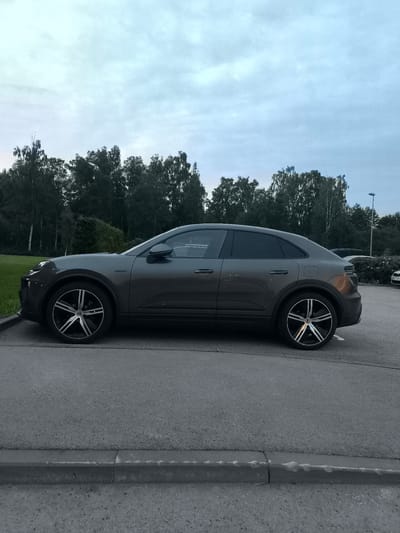 A dark gray Porsche Cayenne coupe is parked on an asphalt lot, viewed from its left side, in Laagri alevik, Estonia. The vehicle is positioned on a paved area next to a grass verge and a line of trees forming a backdrop under an overcast sky. The car features black and silver alloy wheels. In the background, other vehicles and a lamppost are visible. The scene appears to be a public parking area, possibly near a commercial or residential zone.