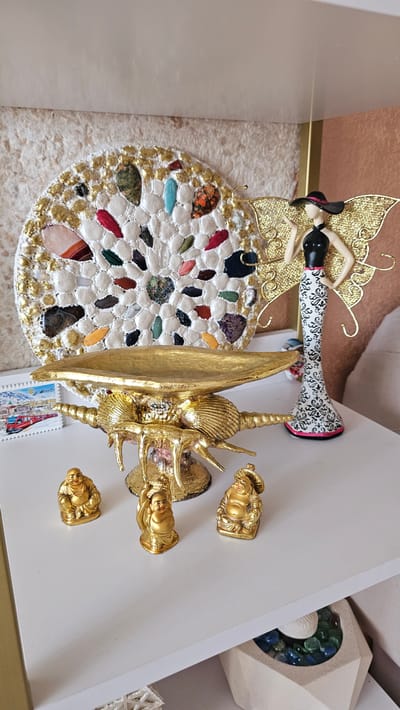 The image displays a collection of decorative items arranged on a white shelf against a textured wall. In the background, a circular mosaic artwork adorned with various colored stones and gold accents is visible. To the right of the mosaic is a figurine of a woman with golden, butterfly-like wings, wearing a black top and a black hat, and a black and white patterned dress.
In the foreground, a golden decorative bowl with an ornate, shell-inspired base rests on the shelf. In front of the bowl, three small golden figurines of what appear to be laughing Buddhas are placed. To the left of the bowl, a small framed picture with a train and a snowy landscape is visible. The shelf is part of a display within an indoor environment. The location is identified as Radkevshchina, Russia.