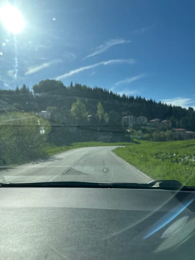 The image is taken from inside a vehicle, looking out the windshield. A road winds uphill through a green, grassy area on the right. Across the road on the left is a tree-covered hillside with buildings visible among the trees. The sky is blue with wispy clouds and the sun is shining brightly in the lower left corner of the frame. The location is Stoikite, Bulgaria.