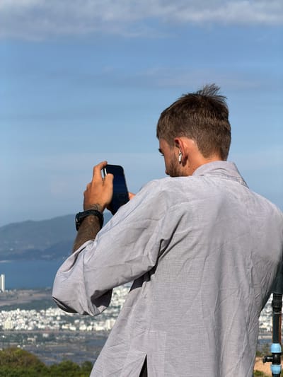 A man in a gray shirt and Airpods is looking at his phone against a backdrop of a blue sky, mountains, and a city. The city, Diên Lạc, Vietnam, is situated along a coastal plain with buildings clustered together. There are trees visible to the right. The man has a wrist watch and a tattoo on his left arm.