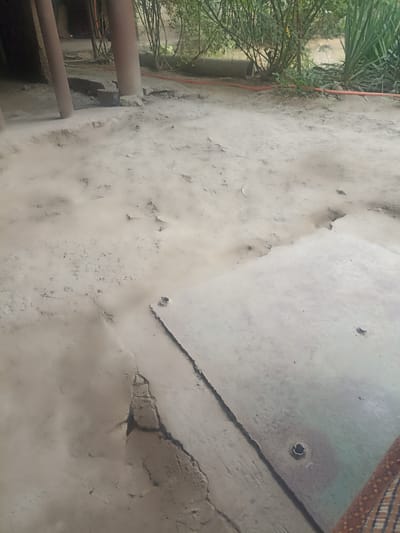 The image depicts an outdoor scene in Aygezard, Armenia, characterized by a rough, dusty ground surface. In the foreground, a cracked, light-colored concrete slab is visible, with a circular hole in its center. To the left of the slab, the ground appears to be composed of packed dirt and dust, with visible imperfections and unevenness. In the background, two large, cylindrical, reddish-brown posts stand vertically, partially obscuring a dense growth of green foliage and trees. A red hose snakes across the ground behind the posts, disappearing into the vegetation. The overall impression is of an unkempt, possibly industrial or construction-related area.