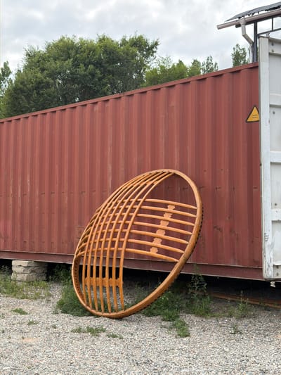 A large, reddish-brown shipping container sits on a gravel surface. Leaning against the container is a large, circular, wooden structure composed of numerous curved slats. The structure appears to be incomplete or possibly a sculptural element. Behind the container, lush green trees are visible against a cloudy sky. To the right of the container, a white section of another structure is present, with a yellow warning triangle sticker that reads "SUPER HEAVY". The ground is covered with small, light-colored pebbles. Some patches of green vegetation are growing in the gravel and underneath the shipping container. The scene appears to be outdoors, likely in an industrial or open area.