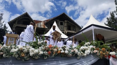 The image shows a group of people gathered in front of a large house in the city of Bukavu, Democratic Republic of Congo. The house is made of stone and has a large, white tent in front of it. The people are all dressed in white robes, and some are holding staffs. The man in the center of the image is holding a leopard skin. There are flowers in front of the people, and the sky is blue with some white clouds. The location, based on coordinates, is likely a place of significance to the community. The scene is possibly a ceremony or celebration. The people appear to be participating in a ritualistic activity or religious practice.