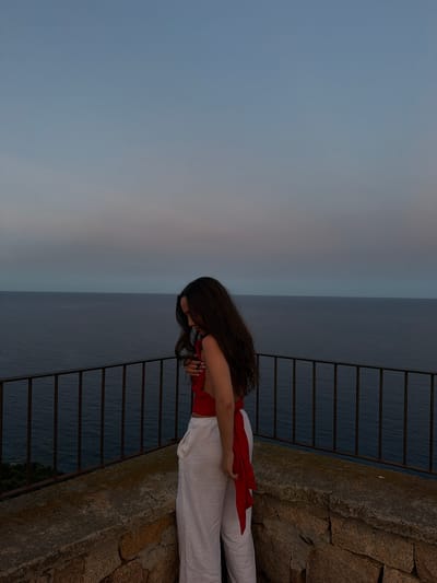 A young woman with long, dark, wavy hair stands on a stone ledge overlooking the sea in Santa Maria Navarrese, Italy. She is wearing a red top and white linen pants. The sky is a soft gradient of blue and pink, indicating either dawn or dusk. A metal railing runs along the edge of the ledge, separating it from the water below. The sea is calm and dark blue. The overall scene conveys a sense of tranquility and contemplation.