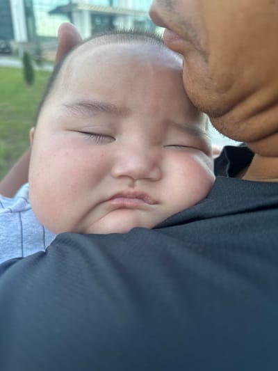 A close-up, vertically oriented photograph shows a person's face gently touching the head of a sleeping child. The child's eyes are closed, and their face is turned towards the viewer, nestled against a dark garment. The adult's chin and jawline are visible in the upper left of the frame, with a hint of stubble. The background appears to be outdoors, with soft, blurred greenery and a building structure visible, suggesting a park or an open area with urban elements. The lighting is soft, indicating it might be daytime, possibly overcast or late afternoon. The overall mood conveyed is one of tenderness and affection. No text is visible in the image. The location is Manas, Kyrgyzstan.
