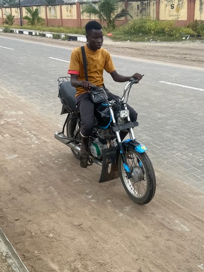 The scene is set in Taupo, Nigeria. A young male, dressed in a mustard yellow t-shirt with red undersleeves and dark trousers, sits astride a motorcycle. He wears dark slide sandals on his feet. A black shoulder bag with a patterned flap is worn across his chest. His hands grasp the chrome handlebars, and he looks slightly to the right of the frame.
The motorcycle features a black seat, a black and blue front fender with yellow and orange accents, spoked wheels, and a square-shaped headlight assembly. An exposed engine block with a green cover is visible on the left side. A black bag is positioned between the handlebars and the rider. A black metal rack is mounted over the rear of the motorcycle.
The motorcycle is positioned on a sandy, unpaved shoulder of a road, with tire tracks and imprints visible in the dirt. To its right, a paved road constructed of grey interlocking pavers in a herringbone pattern is visible, marked with a white painted line. A black and white striped curb separates the paved road from a vegetated strip. In the background, a light yellow-brown wall with darker orange-brown sections is visible. Letters and symbols, including "N", "O", "R" (with a rainbow), and "S" (with a sun), are painted on the wall. Green foliage and palm trees are visible behind the wall. The sky is overcast.