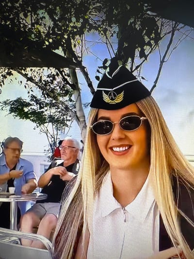The image depicts a woman wearing a black hat with a gold emblem and sunglasses, smiling broadly. She has long blonde hair and is wearing a white collared shirt with a zipper. In the background, two men are seated at a table outdoors, with one wearing a light blue shirt and the other a black shirt. Trees and sky are visible behind them. The scene appears to be a cafe or outdoor seating area. The location is not specified within the image or provided information.