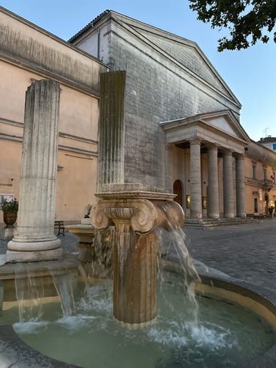 In Anduze, France, in front of a large stone building with a neoclassical facade featuring a portico with columns, a fountain is in operation. The fountain is composed of at least two fluted columns, one of which is topped with a volute-decorated capital, from which water is cascading into a basin. The building appears to be a public institution, as indicated by a sign reading "HOTEL DE VILLE" (City Hall) on an adjacent structure. Two large, fluted stone columns stand prominently in the foreground to the left of the fountain. The scene is set in a public square paved with cobblestones, and a tree branch with green leaves is visible in the upper right corner, suggesting an outdoor setting during daylight hours.